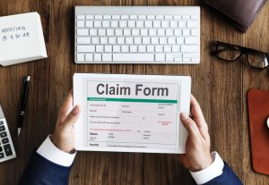 A person holding a tablet showing a generic claim form above a wooden desk with a keyboard and glasses